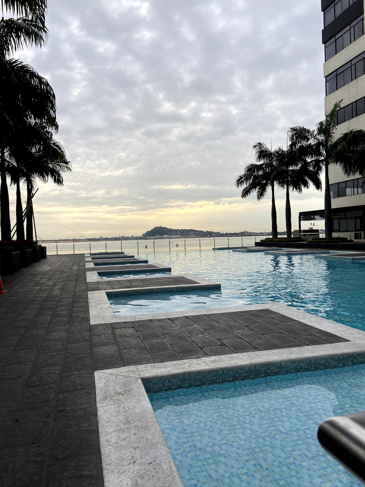 An inviting outdoor pool area features several sunken loungers alongside the water's edge. Towering palm trees frame the view of the calm water and distant hills, while the sky showcases a soft gradient of colors as the sun sets.