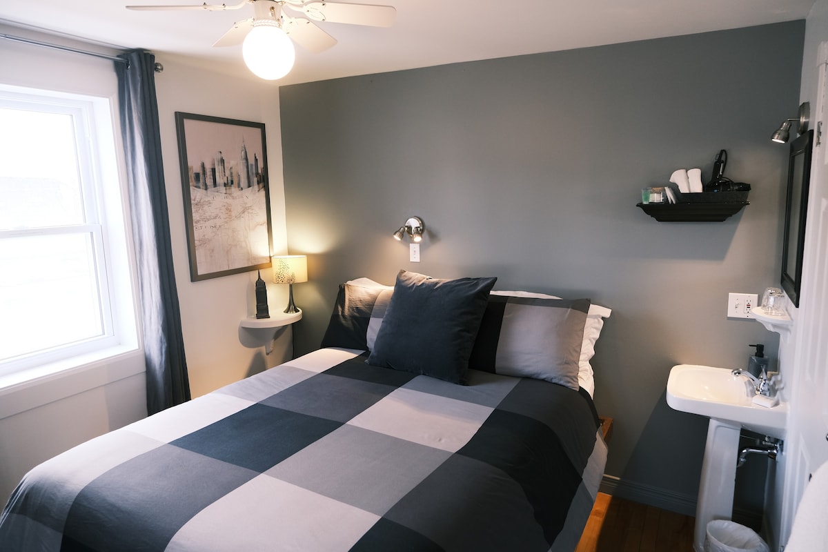 A cozy guestroom features a spacious bed with a checkered gray and white blanket, accented by dark pillows. A small shelf displays toiletries beside a compact sink. Natural light enters through a large window, complemented by gray curtains and a contemporary wall painting.