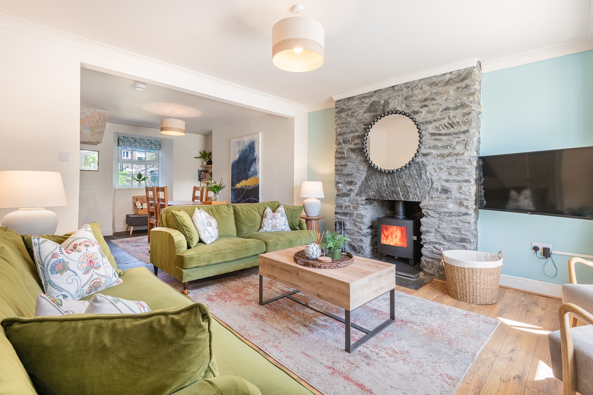 A spacious living room features green sofas and a wooden coffee table at its center. A Lakeland stone fireplace, with a mirror above, adds character to the room. Light streams in from the nearby window, illuminating the warm wooden flooring.