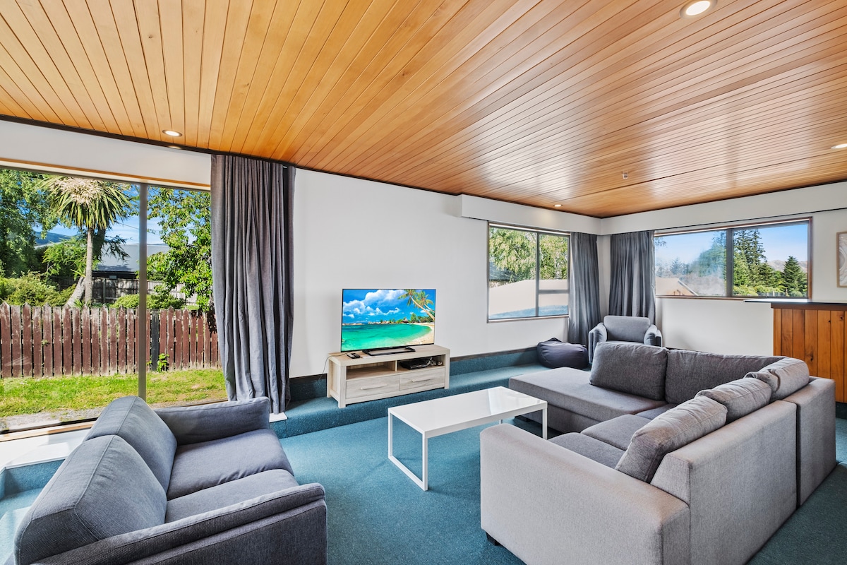 An inviting living area is presented, featuring a large flat-screen television on a light wooden stand. Two gray sofas and a white coffee table create a comfortable seating arrangement, while large windows allow natural light to flow in, showcasing views of the outdoor greenery.