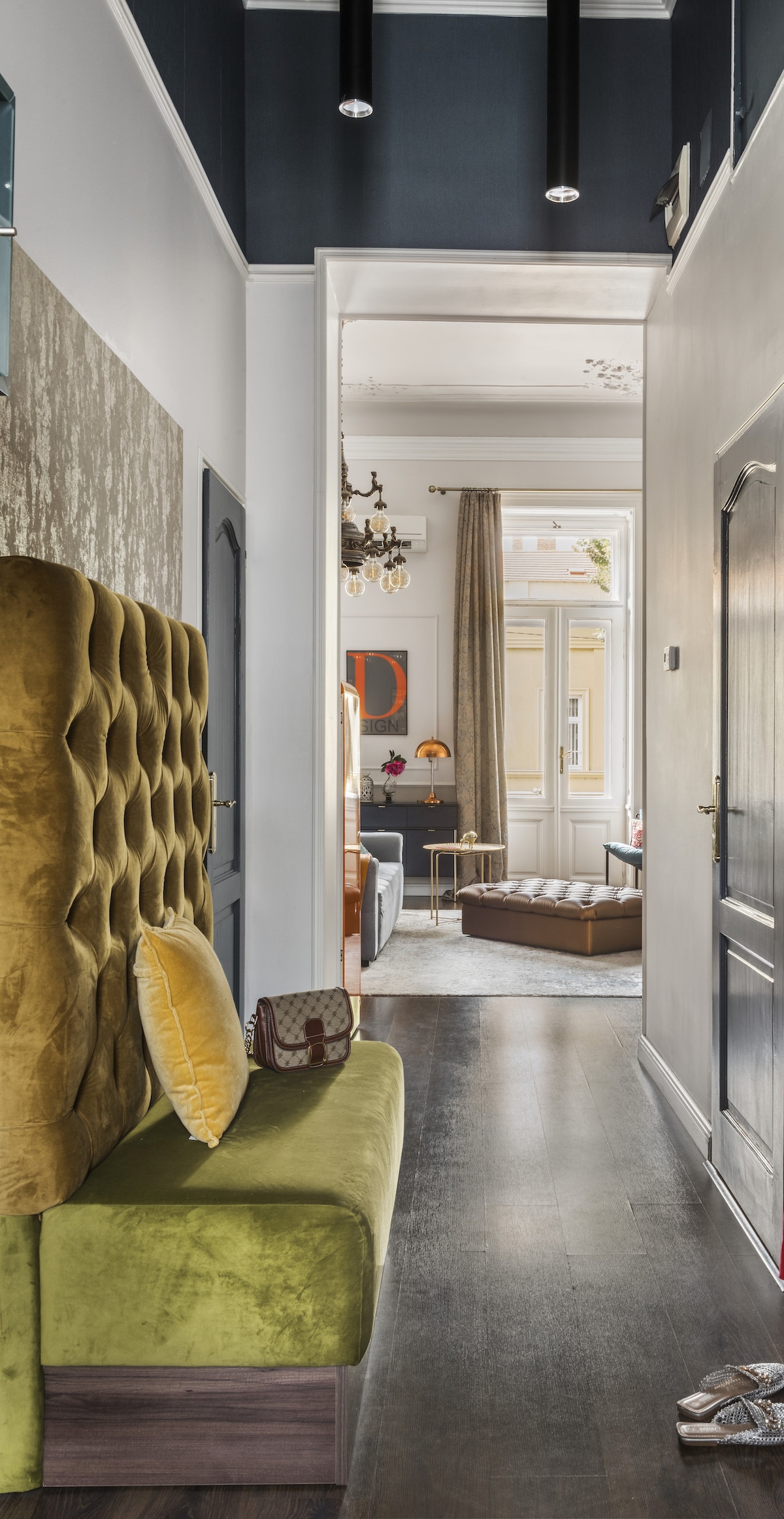 A hall with a plush, green upholstered bench is displayed, providing a welcoming entrance. The space is illuminated by recessed lighting and extends into a well-designed living area, where soft natural light enters through large windows, enhancing the contemporary decor.