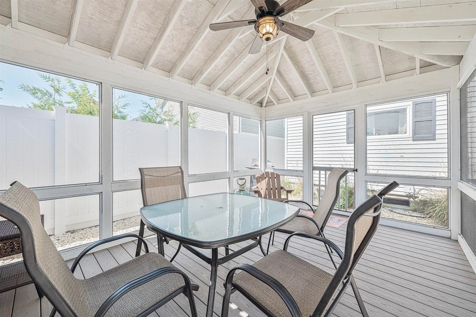 A screened porch is featured with ample natural light. A glass-top table is surrounded by four metal chairs, and a pair of Adirondack chairs is placed in the corner. The ceiling is paneled with exposed beams and a ceiling fan provides ventilation.