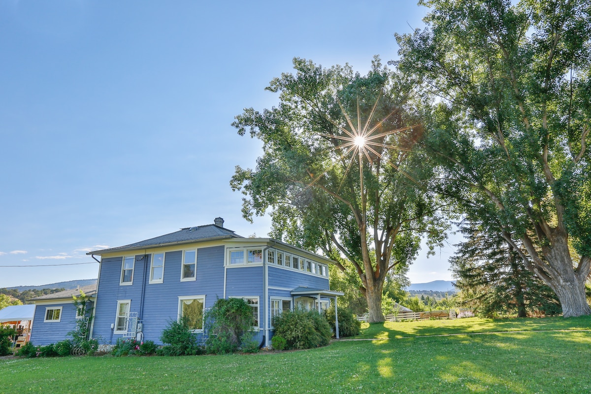 A classic two-story farmhouse, painted in a soft blue, is surrounded by a lush green lawn. Large windows reflect the sunlight streaming through a nearby tree, while distant mountains frame the serene landscape.