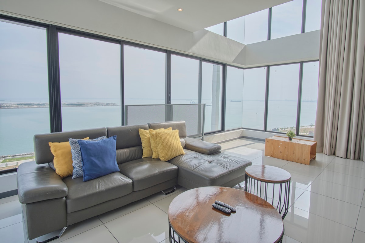 A spacious living area features a modern gray sofa with colorful pillows, offering a tranquil sea view through floor-to-ceiling windows. Round wooden tables are positioned nearby, and light colors create an open atmosphere. Soft curtains are drawn to the side, enhancing natural light.