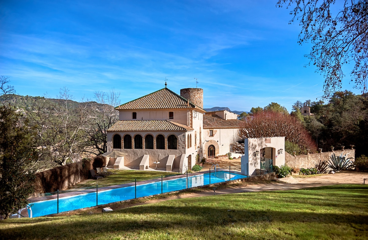 A historic masía is set against a clear blue sky, featuring a well-maintained swimming pool that stretches along the property. The structure showcases a combination of traditional architecture with a prominent tower and a spacious outdoor area surrounded by lush greenery.