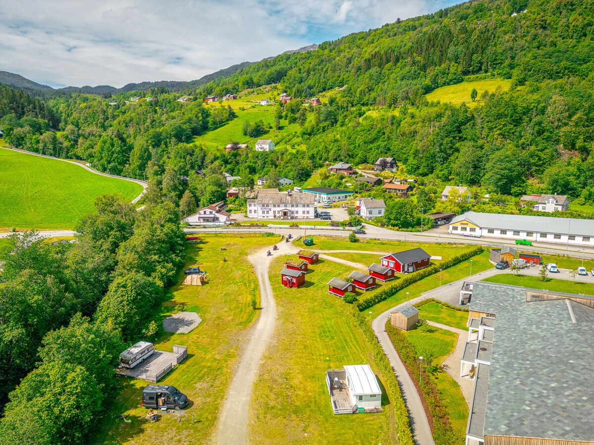 Aerial view of a picturesque landscape featuring a cluster of red huts among lush green fields and trees. A winding road is visible, alongside a small service building, in proximity to a residential area with multiple structures nestled in the hills.