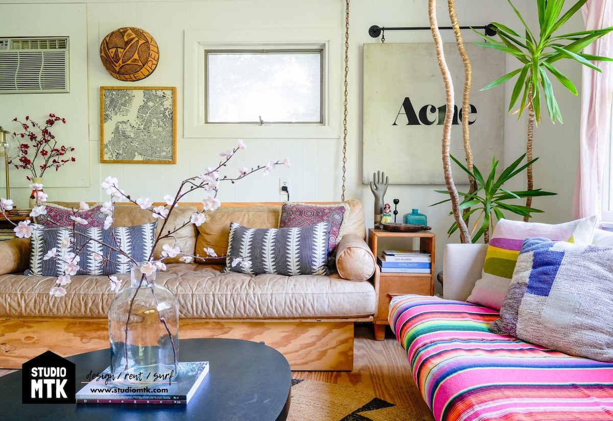 A bright living area features a tan leather sofa adorned with decorative pillows. A colorful striped lounge chair complements a round black coffee table. Potted plants and art pieces add character, while natural light streams through a window in the background.