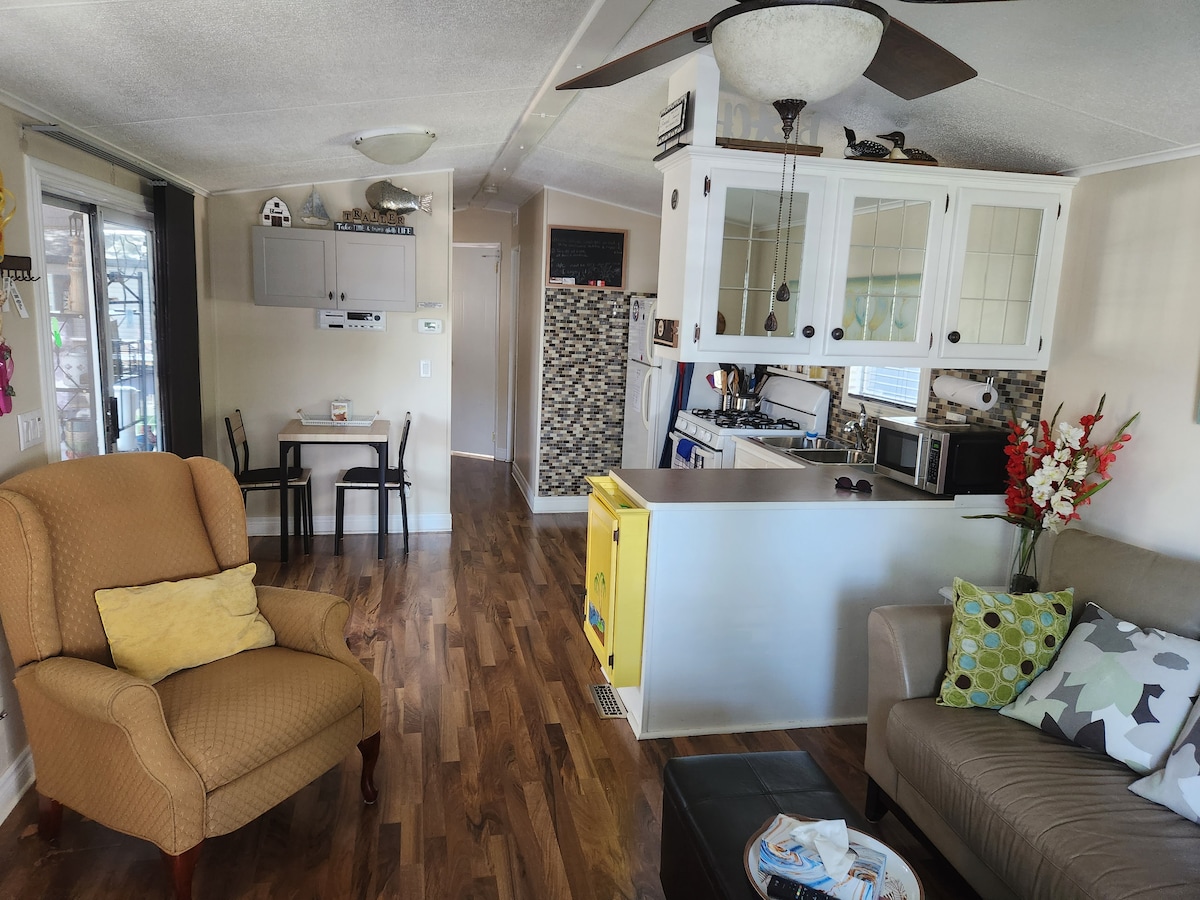 An open living space features a cozy seating area with a brown armchair and a gray sofa, adorned with colorful cushions. A dining table for two is positioned near the kitchen, which is equipped with modern appliances and a decorative backsplash.