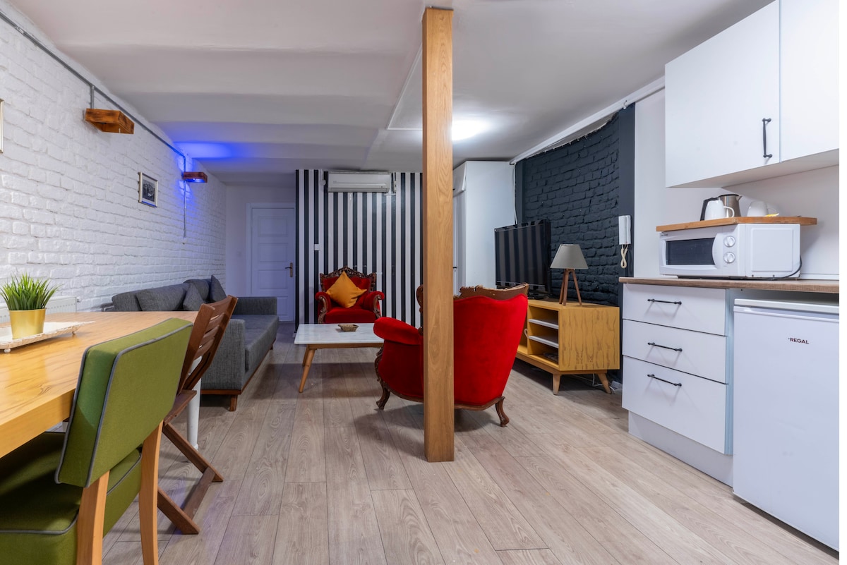 An open-plan living area features a cozy seating arrangement with a gray sofa and a red armchair. A wooden dining table with green chairs is positioned nearby. Bright white walls contrast with dark accent walls, while modern appliances are visible in the well-equipped kitchen.