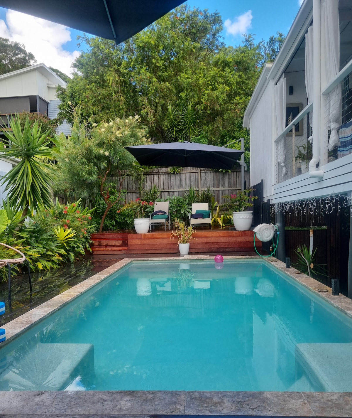 A serene swimming pool is surrounded by lush greenery and vibrant plants. Two lounge chairs are positioned under a large umbrella, creating a relaxing spot for guests. The tranquil atmosphere is enhanced by wooden decking that leads to the pool area.