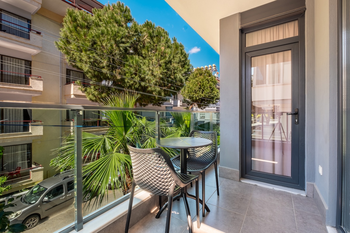 A private balcony features a round table and two chairs, surrounded by lush greenery. The view includes tall trees and palm plants, contributing to a relaxing outdoor space. A glass door leads to the interior, allowing natural light to enhance the atmosphere.