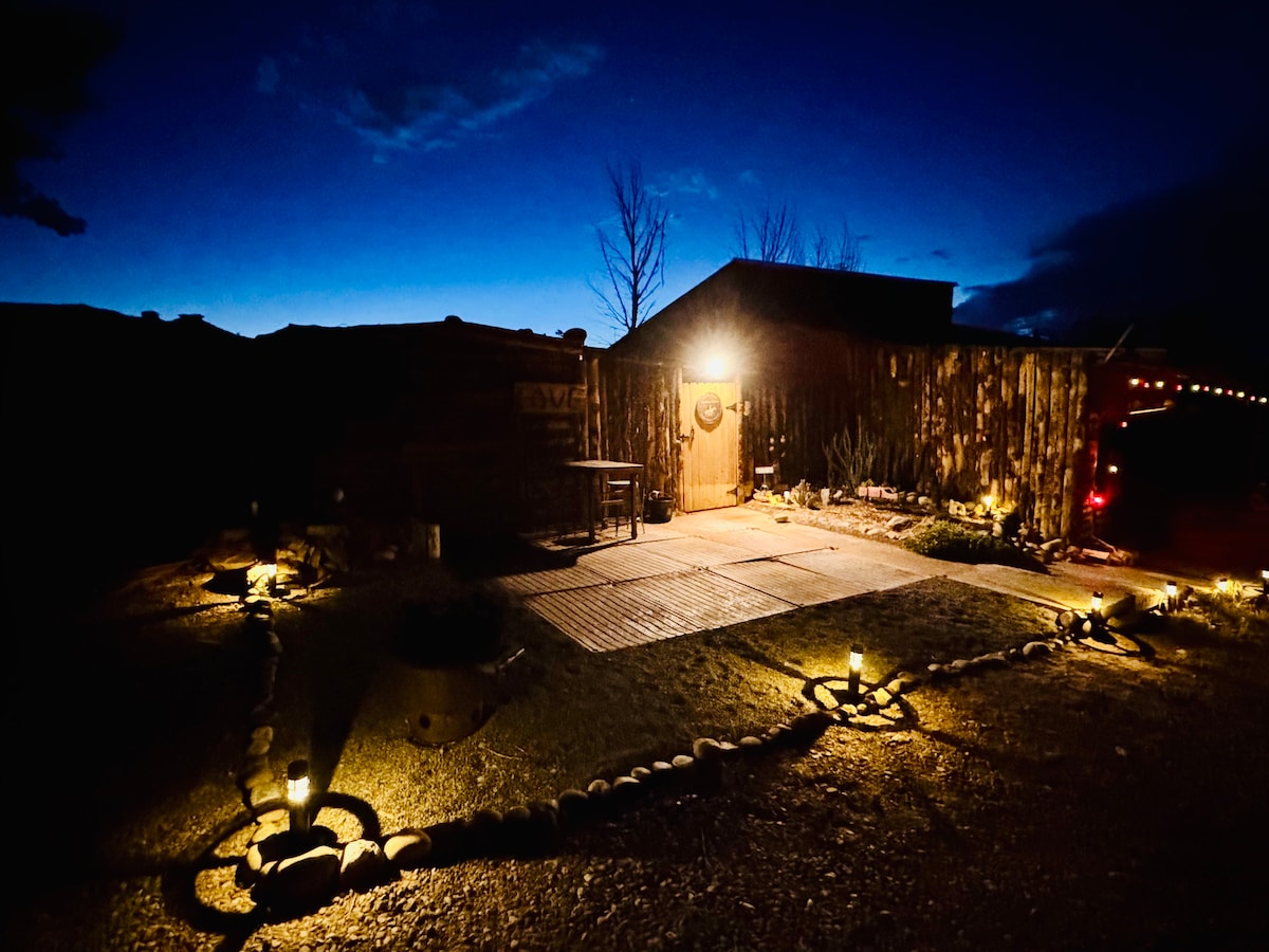 The cabin exterior is softly illuminated at dusk, highlighting its rustic wooden structure and welcoming entrance. Surrounding the path, small lights and stones define the outdoor space, enhancing the tranquil atmosphere of the natural landscape.