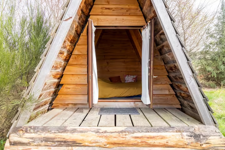 A wooden tipi is featured with an open front, showcasing a spacious sleeping area inside. The entrance includes two white curtains partially drawn back, revealing a cozy mattress with decorative cushions. The wooden deck offers a natural surface leading into the surrounding greenery.
