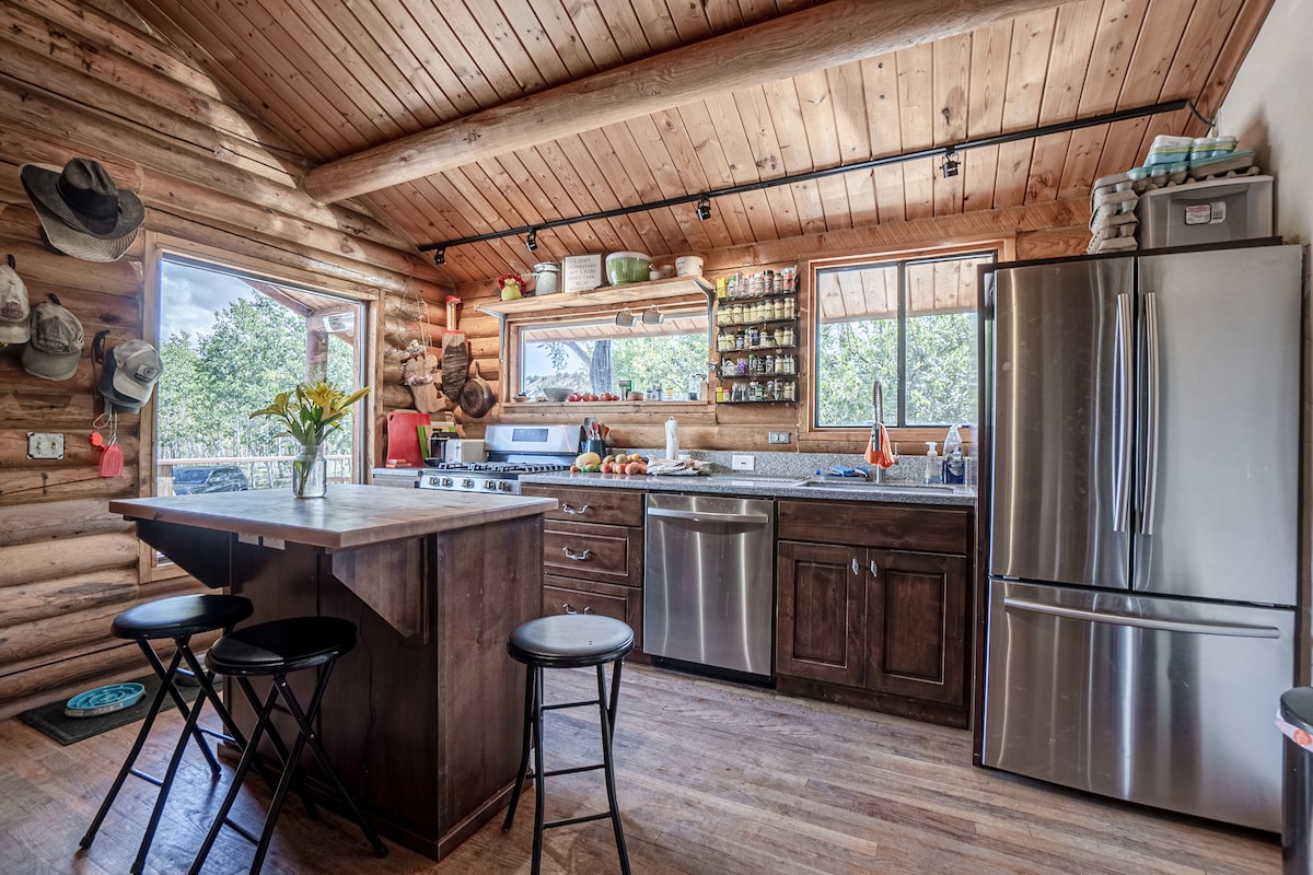 The kitchen features stainless steel appliances and a spacious countertop, complemented by wooden cabinetry. Natural light enters through two windows, enhancing the rustic decor. A central island provides additional seating with several black stools, and fresh flowers are displayed on the counter.