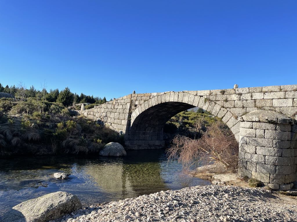 A stone arch bridge spans over clear water, surrounded by natural greenery and rocky terrain. The blue sky above contrasts with the earthy tones of the landscape, highlighting the serene setting. Reflections can be seen on the water's surface, enhancing the scene's tranquility.