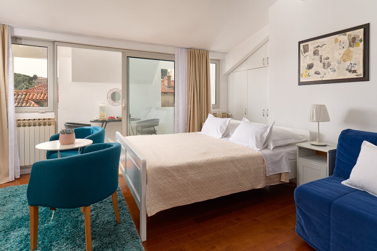 A bright and airy sleeping area features a spacious bed with light bedding. Two chairs with soft, blue upholstery are positioned near a small table. Natural light filters through large windows, illuminating the wooden floor and providing views of the surrounding rooftops.