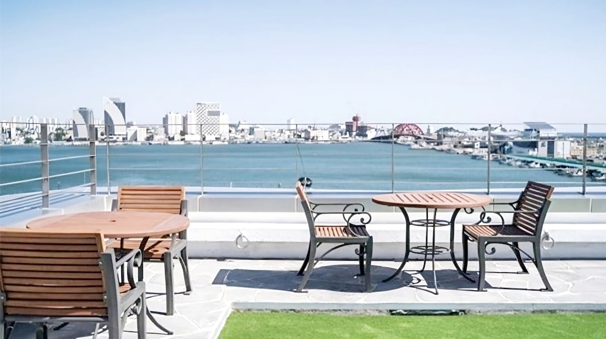 An outdoor terrace features several wooden tables and chairs arranged around a central table, offering a scenic view of the waterfront. The skyline is visible in the background, with a clear blue sky overhead, creating an inviting space for relaxation.