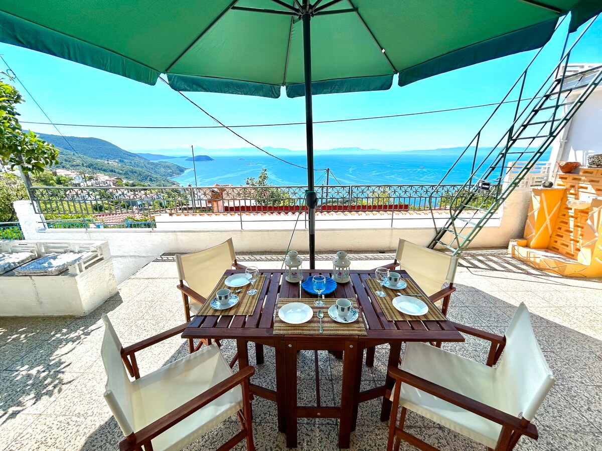An outdoor dining area features a wooden table set with plates and glasses under a large green umbrella. The backdrop reveals a panoramic view of the sea and surrounding landscape, complemented by a clear blue sky. White cushioned chairs surround the table.
