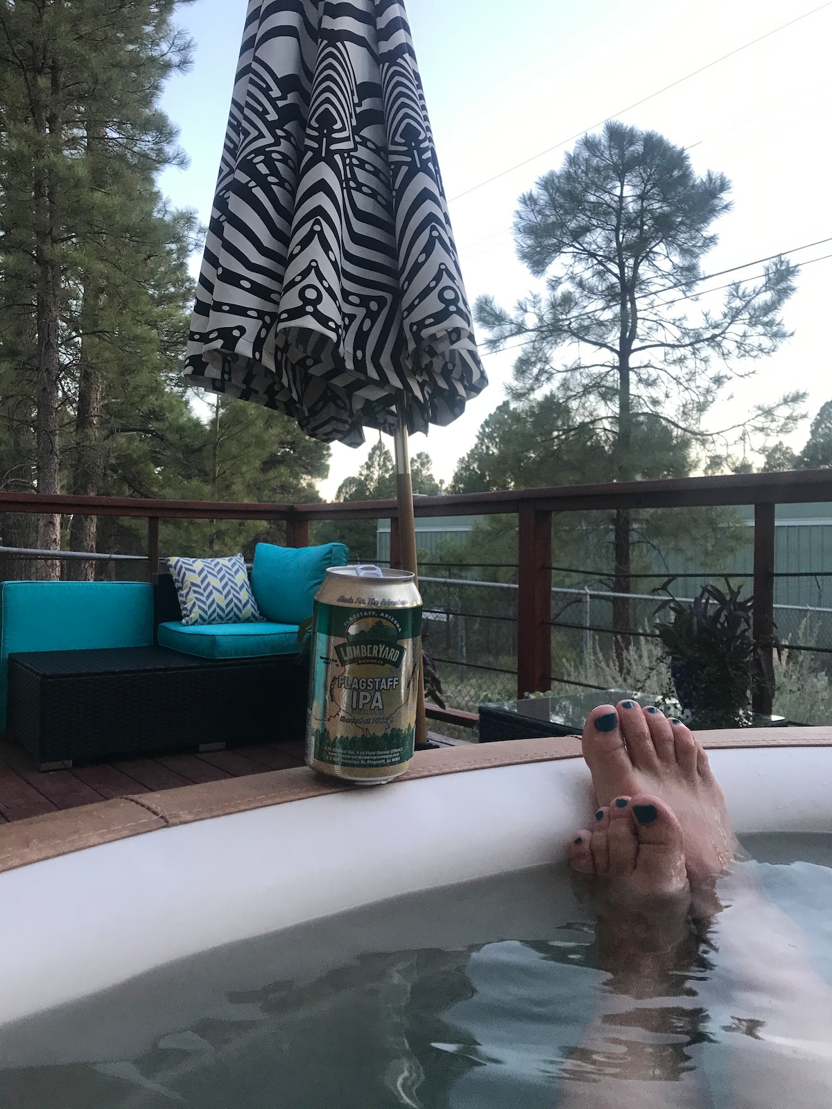 A hot tub is positioned on a wooden deck, surrounded by greenery. A colorful umbrella offers shade, while a can of beer rests nearby. Comfortable seating is visible in the background, enhancing the relaxing ambiance of the outdoor space.