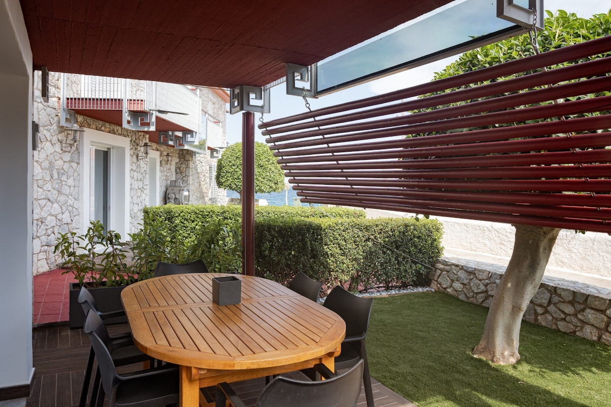 An outdoor dining area features a round wooden table surrounded by six black chairs. Above, horizontal wooden slats provide shade, while a neatly maintained green lawn and landscaped bushes enhance the ambiance. The background reveals views of the neighboring home's stone facade.