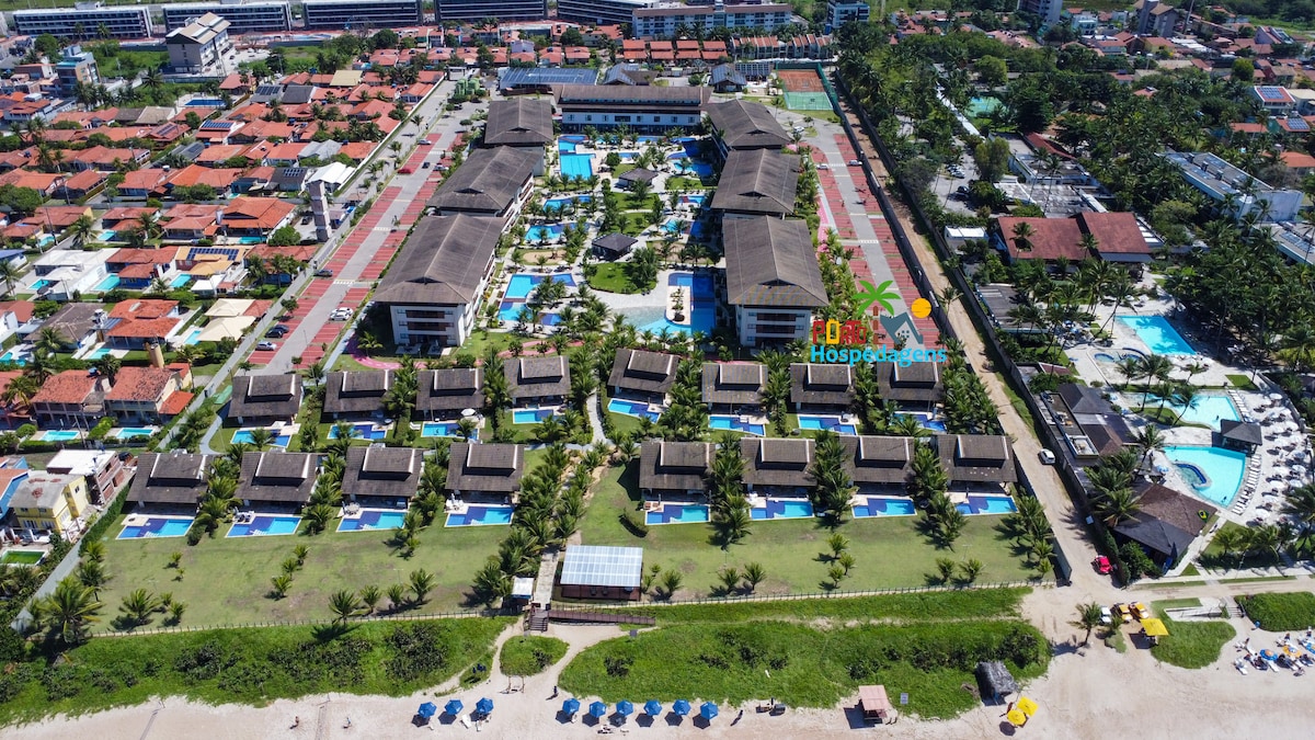An aerial view captures the expansive resort grounds, featuring numerous pools and well-maintained gardens. The layout includes several buildings and bungalows, surrounded by lush greenery. The proximity to the beach is evident, with beach chairs arranged along the sandy shore.