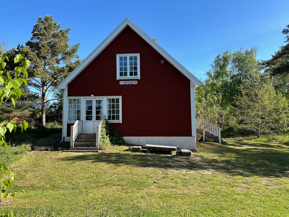 The red wooden cottage is framed by a natural setting, showcasing a pitched roof and large windows. A small set of steps leads to the entrance, while a grassy area surrounds the house, accented by trees and shrubs.