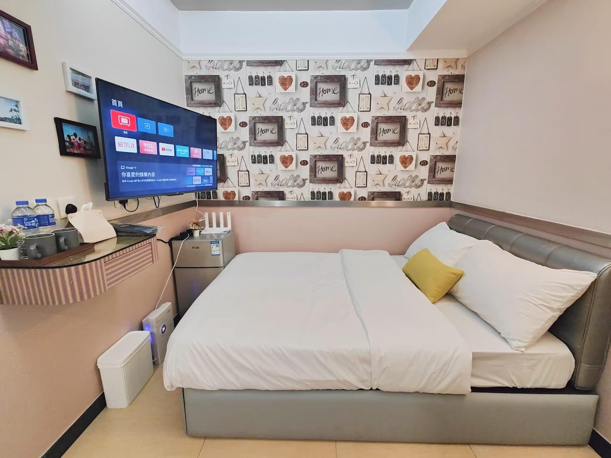 A compact bedroom is displayed, featuring a spacious bed with a light duvet and a decorative cushion. A wall-mounted television provides entertainment options, and a mini-fridge is visible. Shelving with bottled water and toiletries complements the area, enhancing functionality.