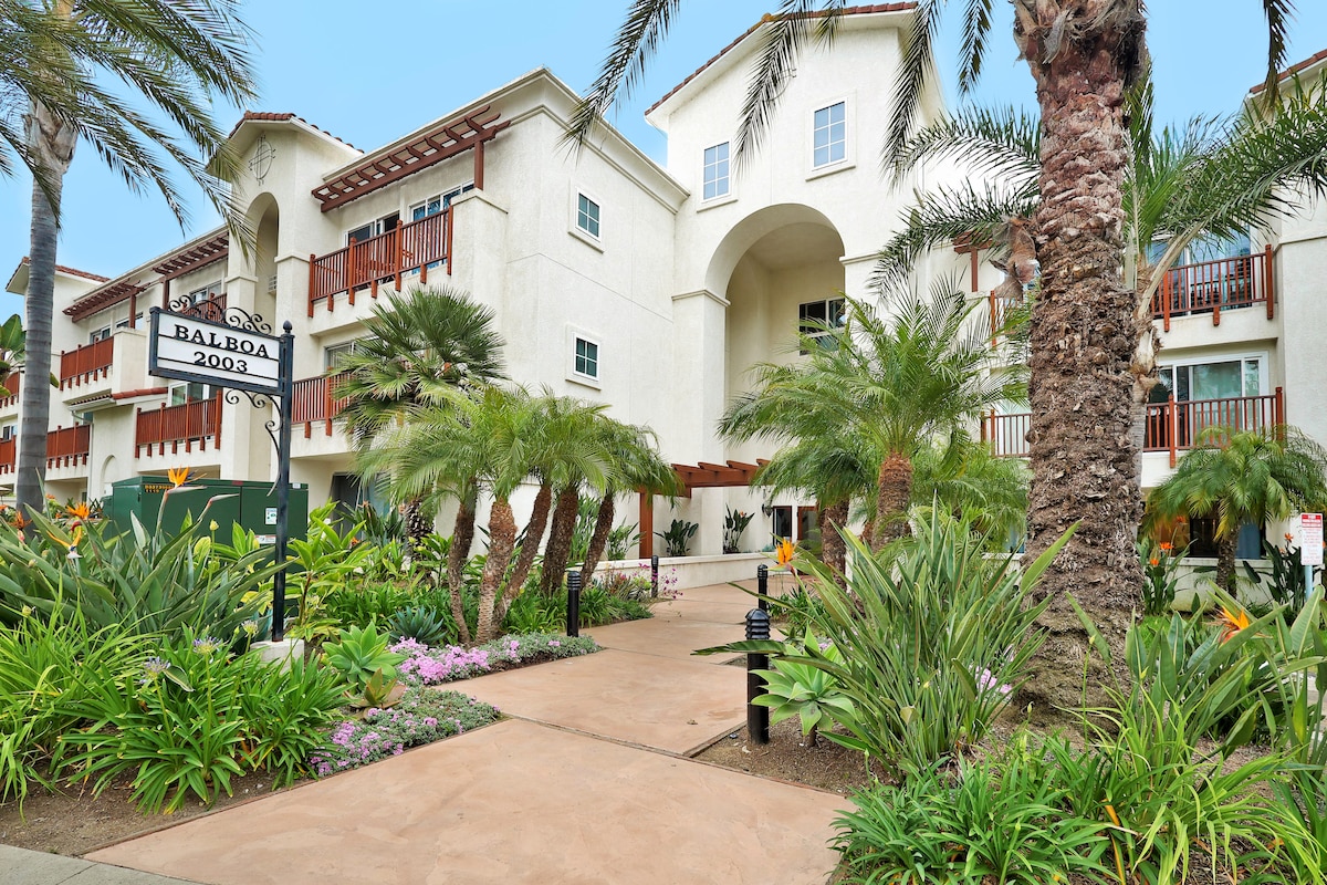The entrance to a condo complex is framed by lush palm trees and vibrant landscaping. Stone pathways lead towards the building, which features a white exterior with terracotta roof accents. A sign indicating 'Balboa 2003' is visible near the entrance, blending harmoniously with the surroundings.