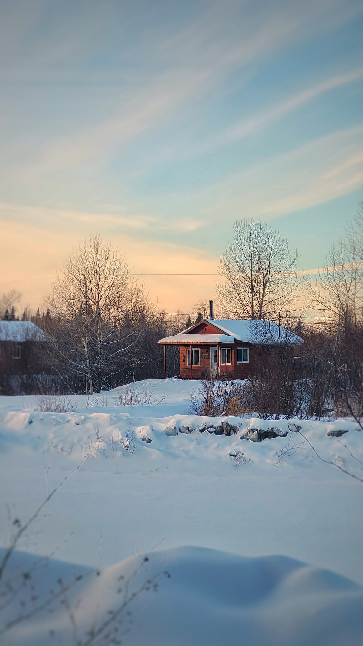 A rustic wooden chalet is positioned amidst a snowy landscape, surrounded by bare trees. The structure features a sloped roof covered in snow, with warm light visible through the windows as evening light casts a gentle hue across the scene.