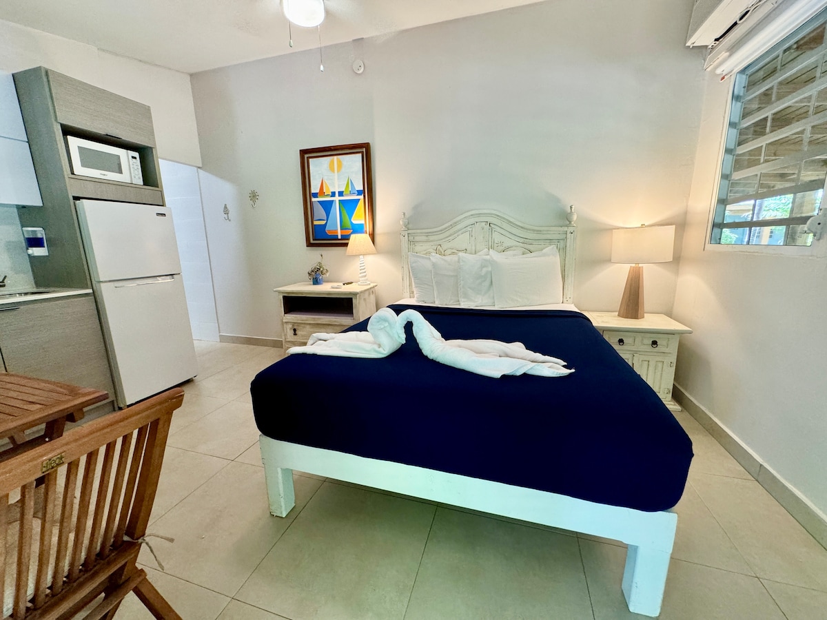 A spacious studio is presented, featuring a comfortable queen-size bed draped with a blue coverlet. Light neutral-toned walls complement wooden furniture, including a small table and chairs. A kitchenette is visible along with a framed art piece and soft lighting near the window.