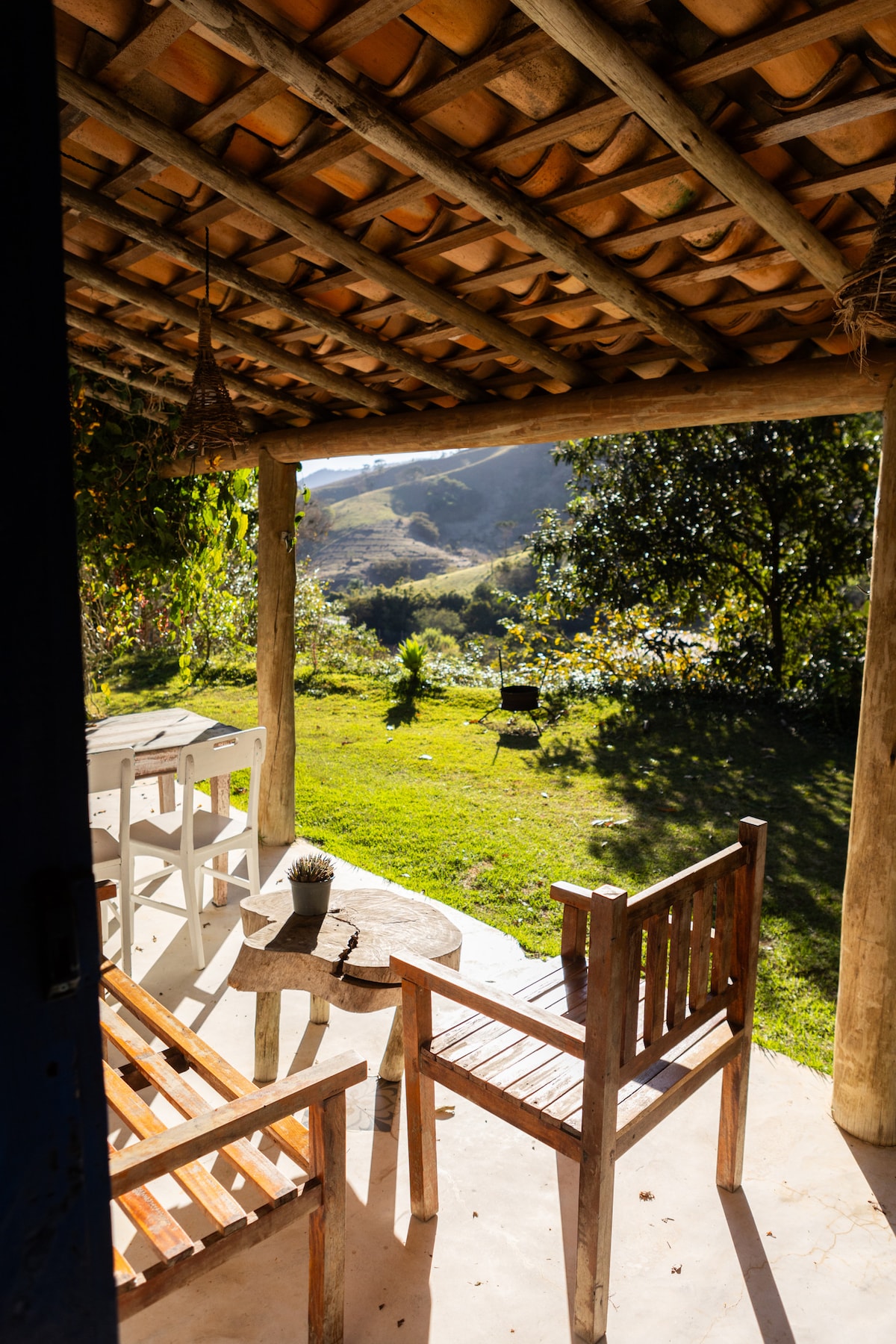 A charming patio area features wooden chairs and a small table, surrounded by vibrant greenery and rolling hills in the distance. The structure has a rustic roof made of wooden beams and thatch, providing a cozy spot to relax and enjoy the natural landscape.