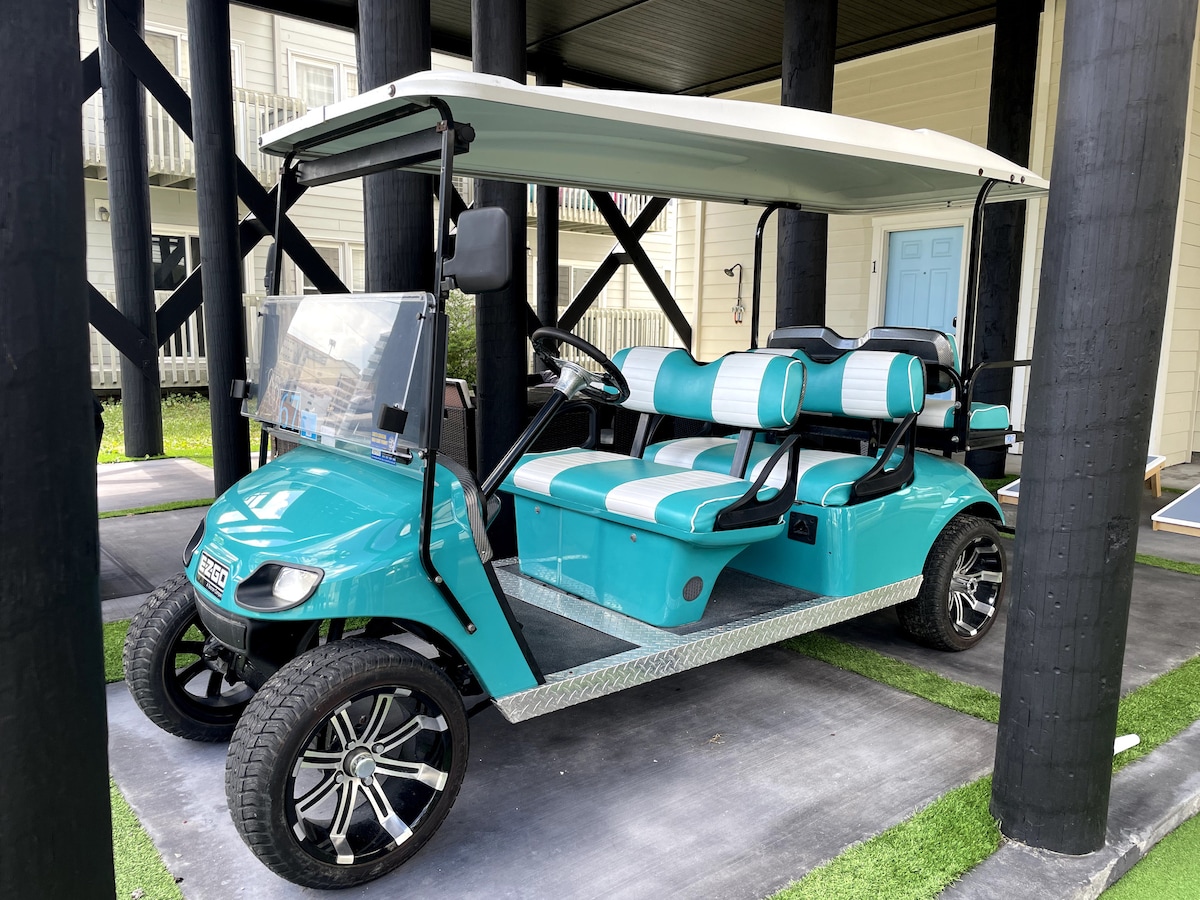 A turquoise golf cart is parked under a covered area, featuring four seats with a teal and white design. The cart has sleek black wheels and a spacious area for storage in the back, providing convenience for travel around the neighborhood.
