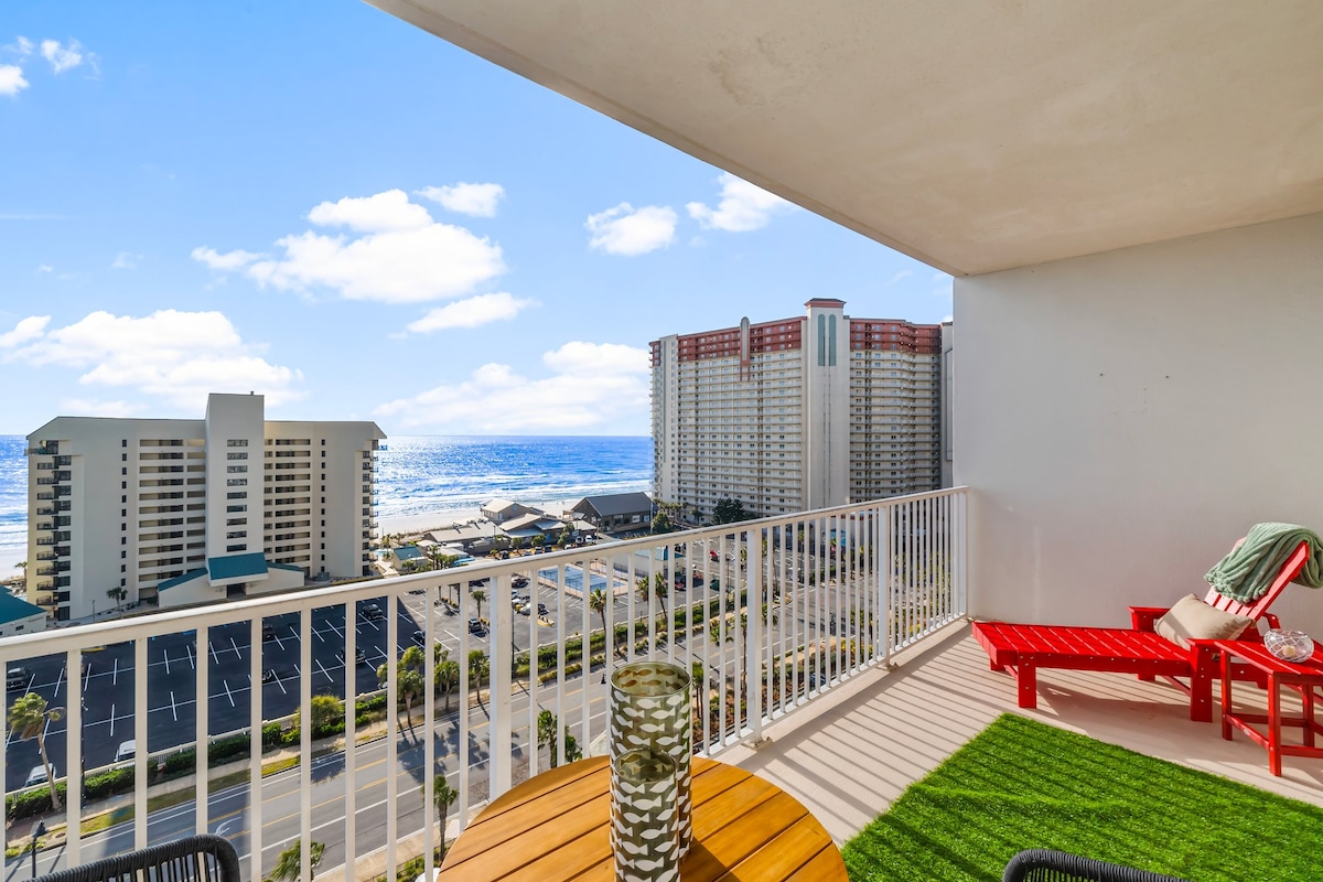A private balcony offers views of the Gulf and nearby buildings, complemented by outdoor seating. A round wooden table is positioned with a tall decorative candle holder on its surface. Bright red lounge chairs and a green area rug add a pop of color to the space.