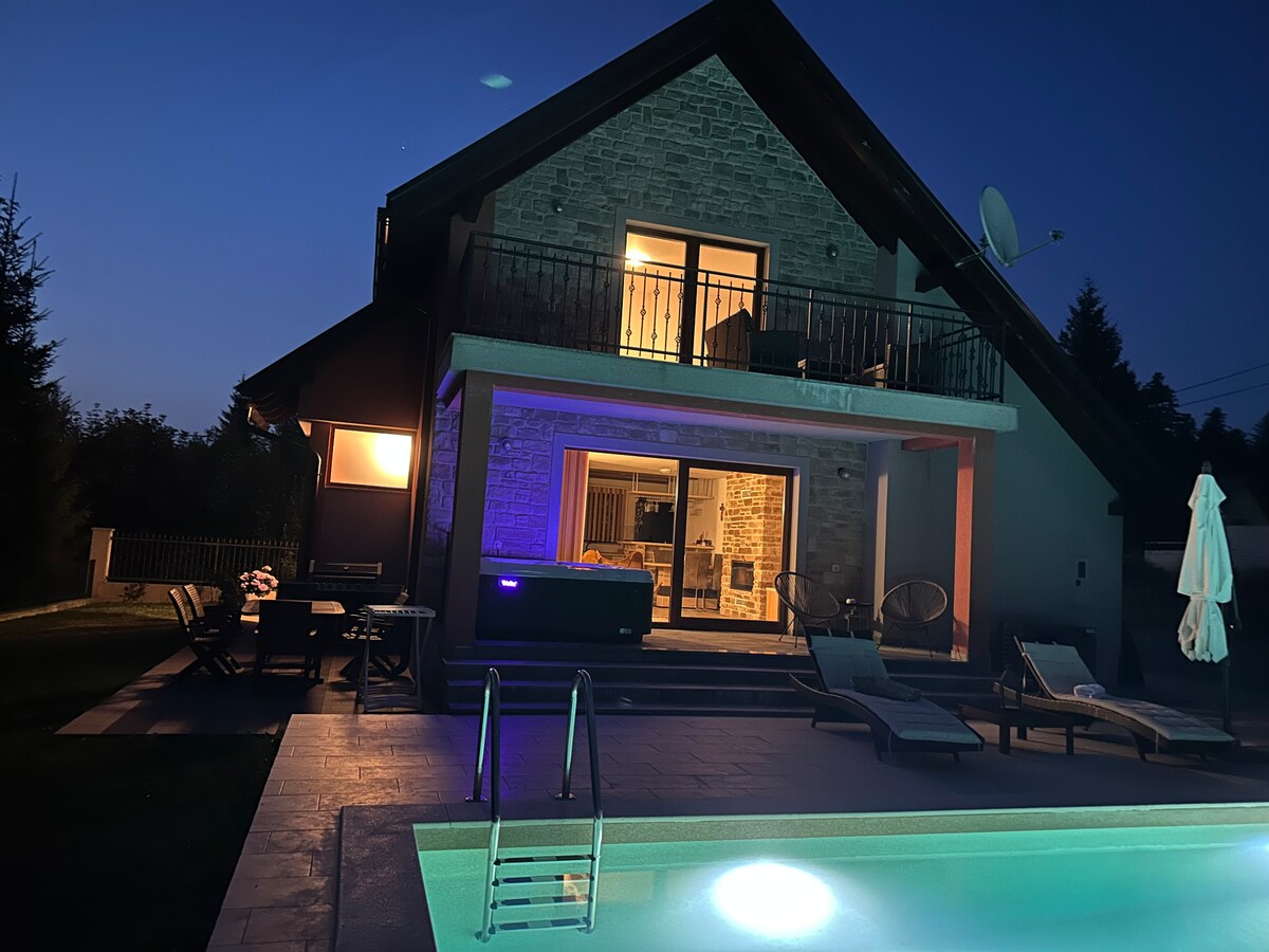 The villa is illuminated at night, showcasing its stone facade and modern architectural features. A heated seasonal pool is visible in the foreground, with lounge chairs arranged nearby. The outdoor massage hot tub and dining area are also evident, creating a serene ambiance.