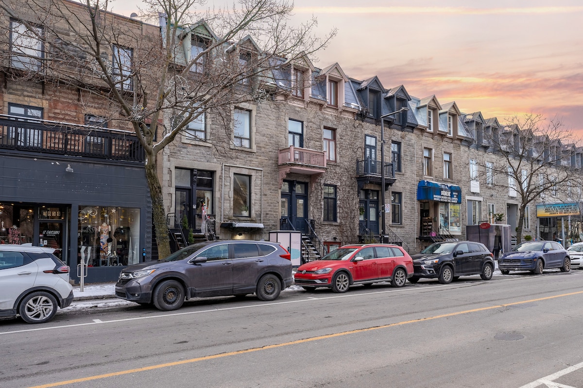 A street view of a classic Montreal building showcases its elegant stone façade and multi-colored roofs. Parked cars line the street, with storefronts visible at ground level. A tree stands beside the building, and the sky displays soft pink and orange hues from the sunset.