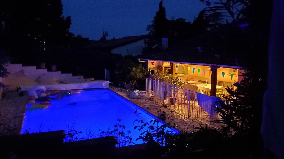 An inviting outdoor space is illuminated by the soft glow of the pool and nearby lights. The swimming pool, surrounded by greenery, features floating lounge items. A covered terrace with festive decorations offers a welcoming area for relaxation, completing the tranquil evening atmosphere.