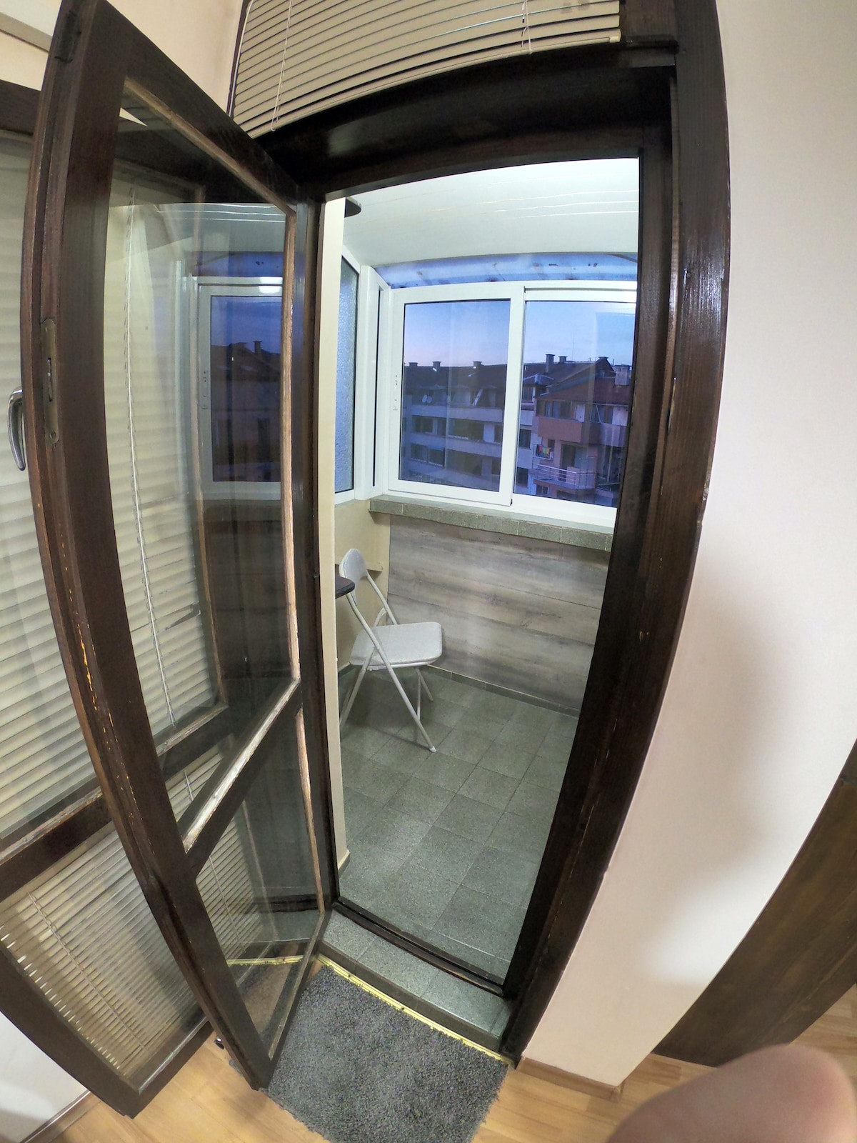 A small balcony is viewed through a glass door, featuring a single chair and large windows that provide a view of the surrounding neighborhood. The floor is tiled, and light wooden accents enhance the space's modern feel.