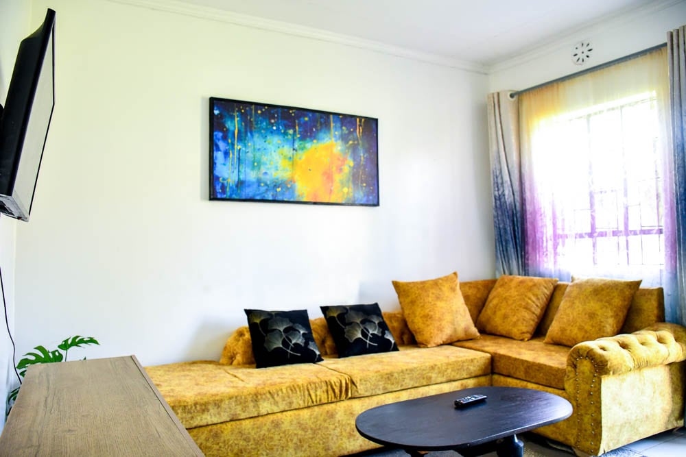A living area features a light-colored sectional sofa adorned with dark pillows. A coffee table sits in front, positioned under a colorful piece of wall art. Gentle light enters through a window, complemented by sheer and solid curtains.