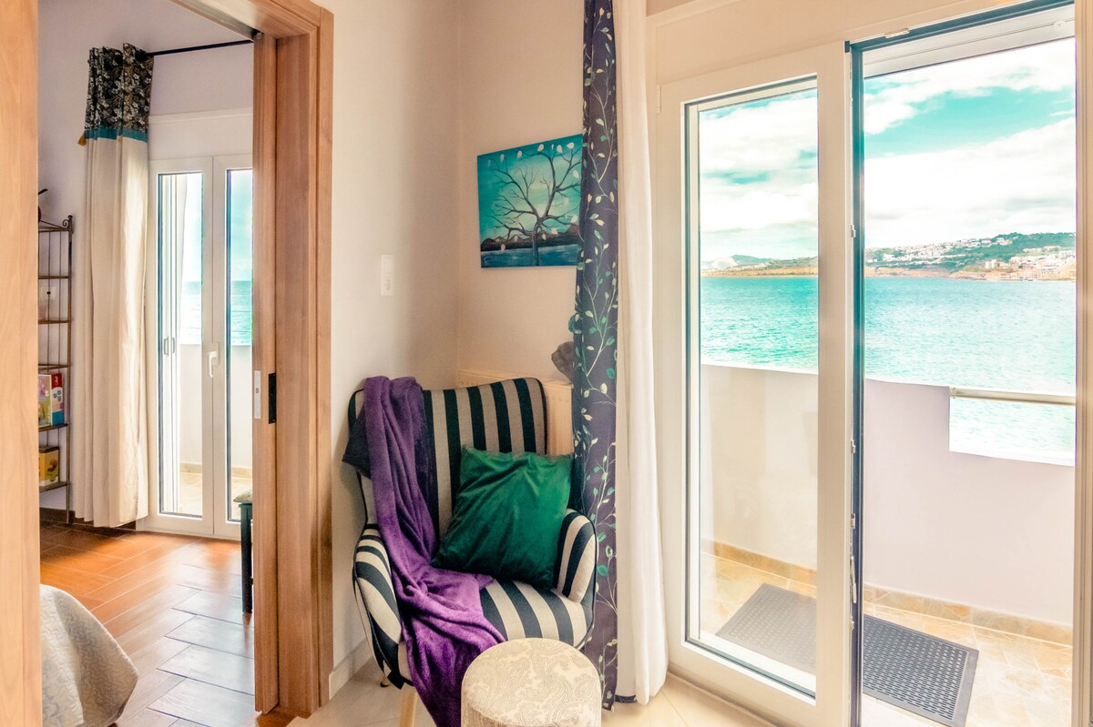 A seating area features a striped armchair draped with a soft purple blanket, complemented by decorative pillows. Large glass doors provide a clear view of the sea and open to a balcony. Natural light fills the space, enhancing the serene atmosphere.