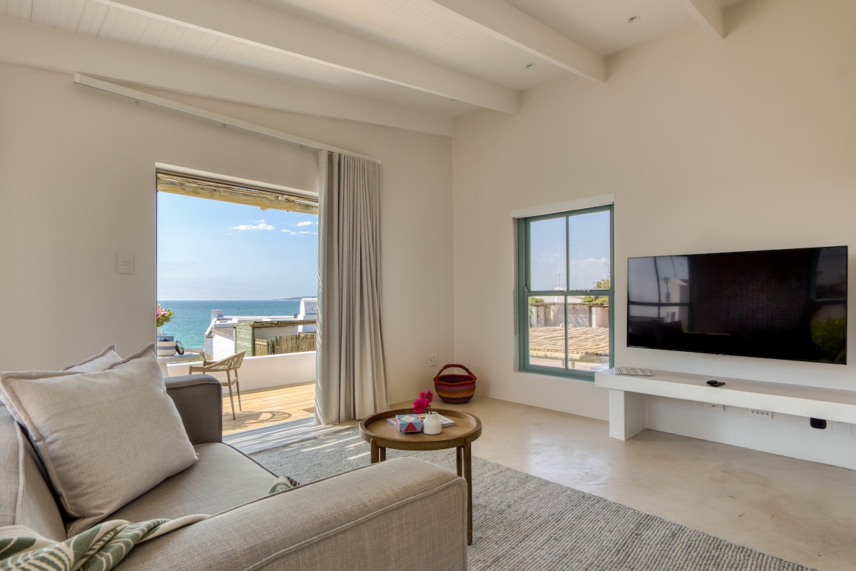 A comfortable living area features a neutral sofa positioned beside a circular coffee table. Large windows provide an unobstructed view of the sea. Sunlight fills the space, highlighting a television mounted on the wall. A small, inviting outdoor space is visible beyond.