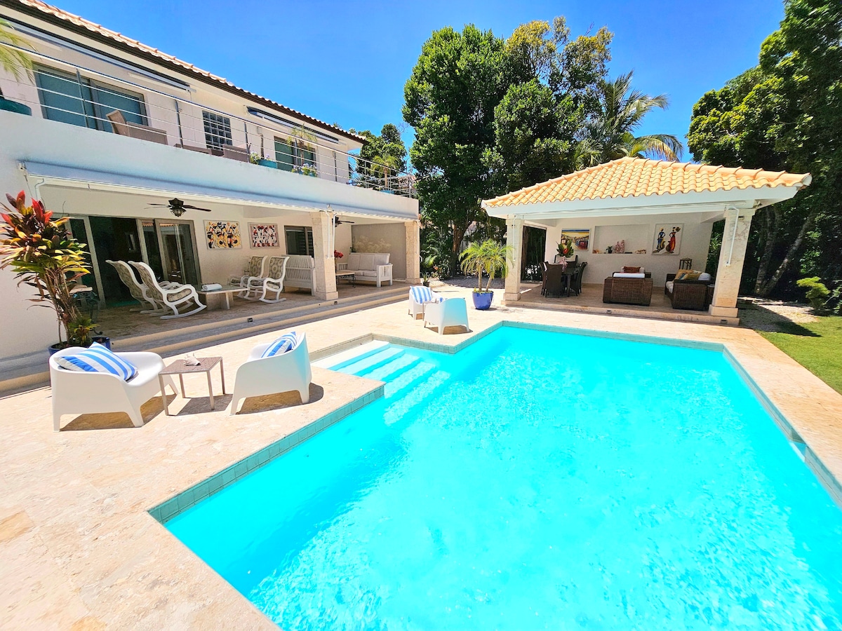 A private pool is surrounded by a spacious terrace, with lounge chairs positioned nearby. A shaded area features outdoor seating, while the villa's two-story structure is visible in the background, framed by lush greenery.