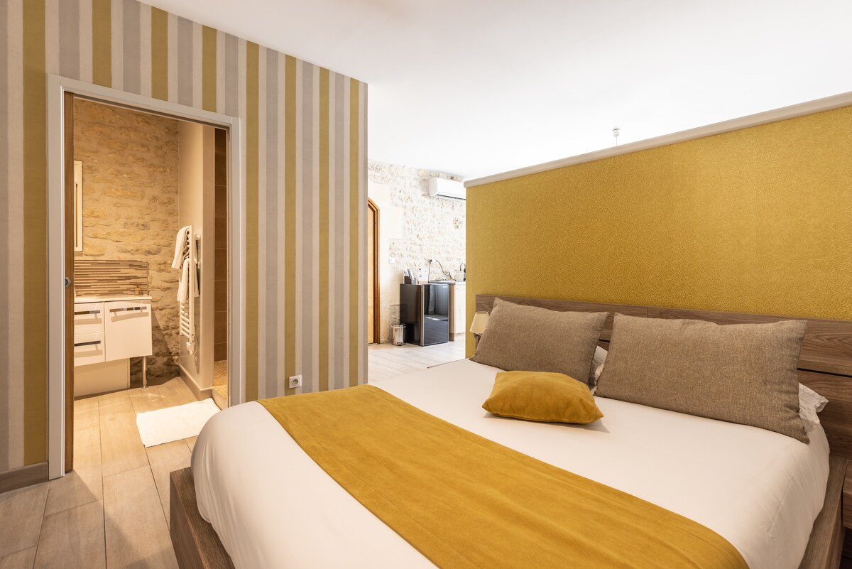 A bright sleeping area features a spacious bed adorned with neutral-toned pillows and a mustard-colored throw. The walls are decorated with subtle stripes, and a doorway reveals a glimpse of a modern bathroom with stone accents.