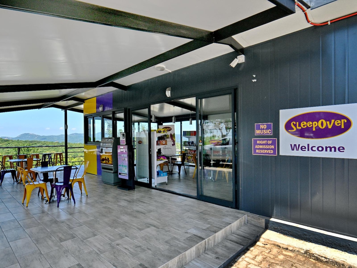 The exterior of the SleepOver KMIA welcome area is shown, featuring a shaded patio with colorful chairs and tables. Large windows allow views of the surrounding landscape, while a welcoming sign highlights the establishment's name. A no music sign indicates a quiet atmosphere.