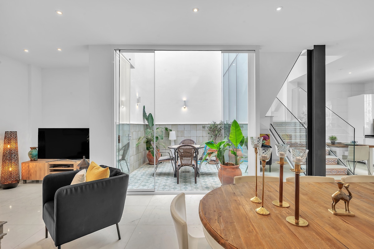 A bright and open living area features a modern dining table with decorative candles and elegant seating. Large sliding glass doors provide a view of a private patio adorned with greenery. Natural light illuminates the space, enhancing the clean lines and contemporary design.