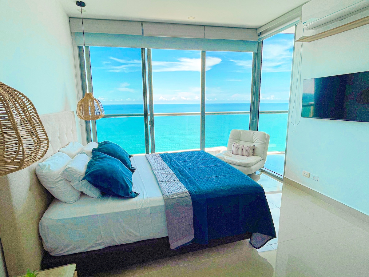 A serene bedroom offers expansive windows showcasing stunning ocean views. A spacious bed with blue and white linens is complemented by a comfortable armchair. A wall-mounted TV is visible, and natural light fills the room, enhancing the tranquil ambiance.
