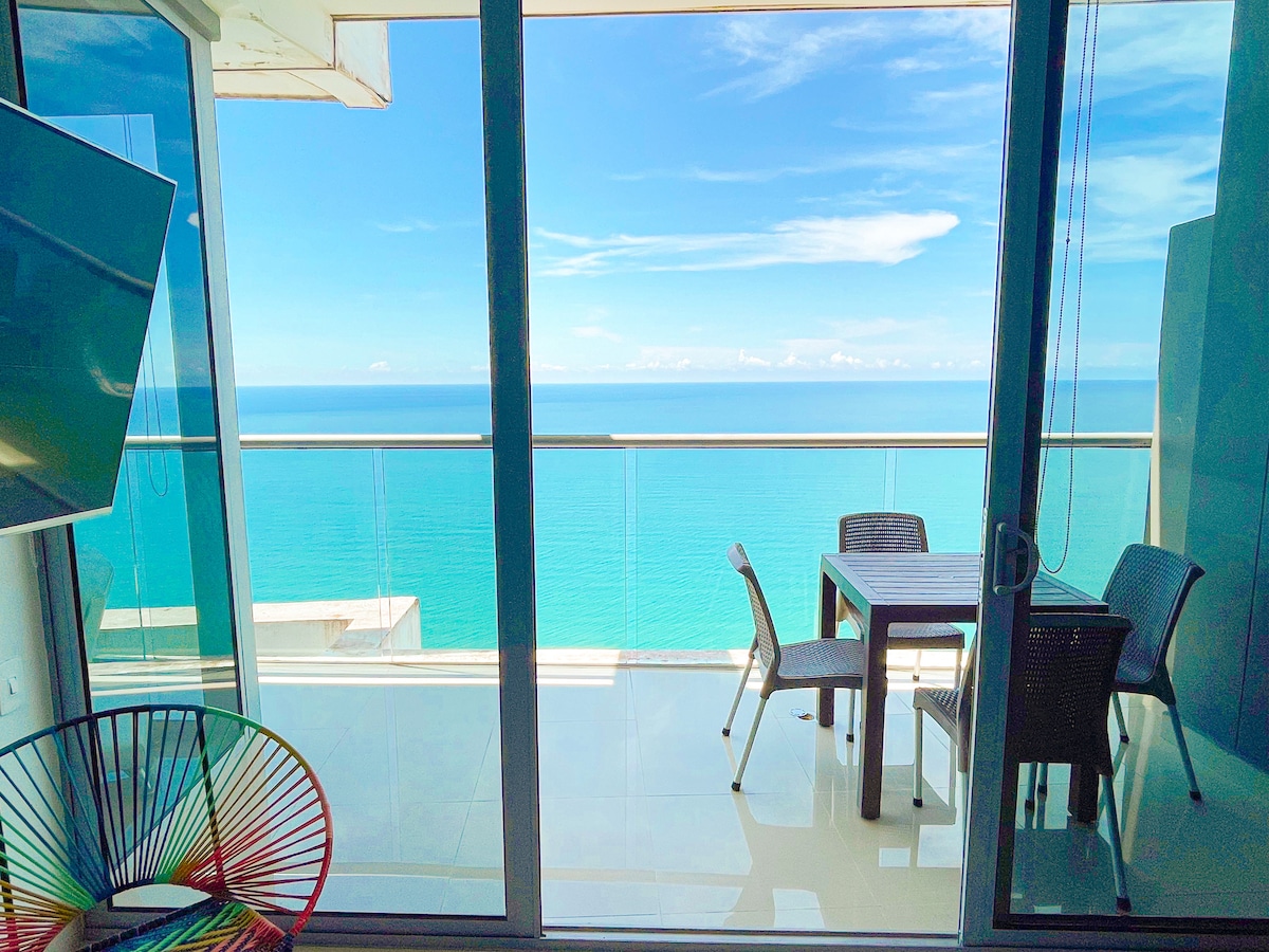 A panoramic ocean view is presented from the balcony, where a table and chairs create a casual dining setting. The interior features large glass doors that invite natural light into the space, highlighting the serene blue hues of the water beyond.