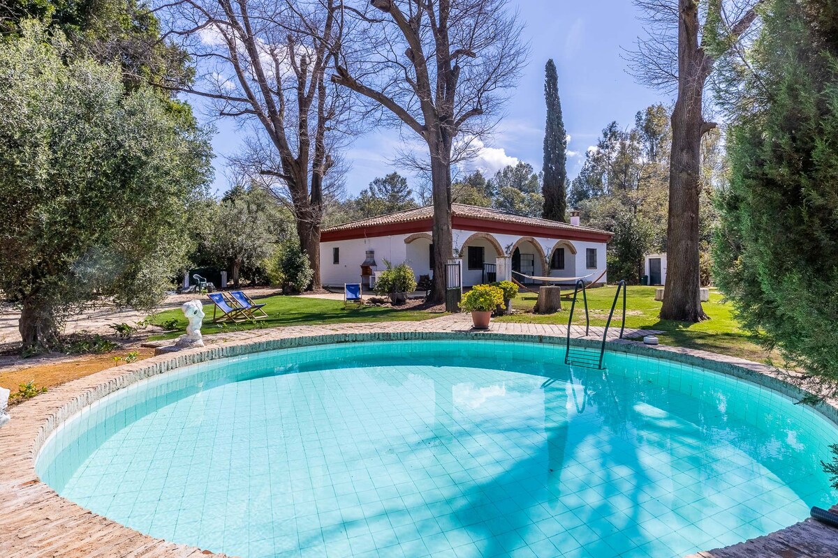 A circular pool with clear turquoise water is framed by lush greenery and large trees. Lounge chairs are positioned nearby, inviting relaxation. The white house in the background features arched windows and is surrounded by an expansive garden space.