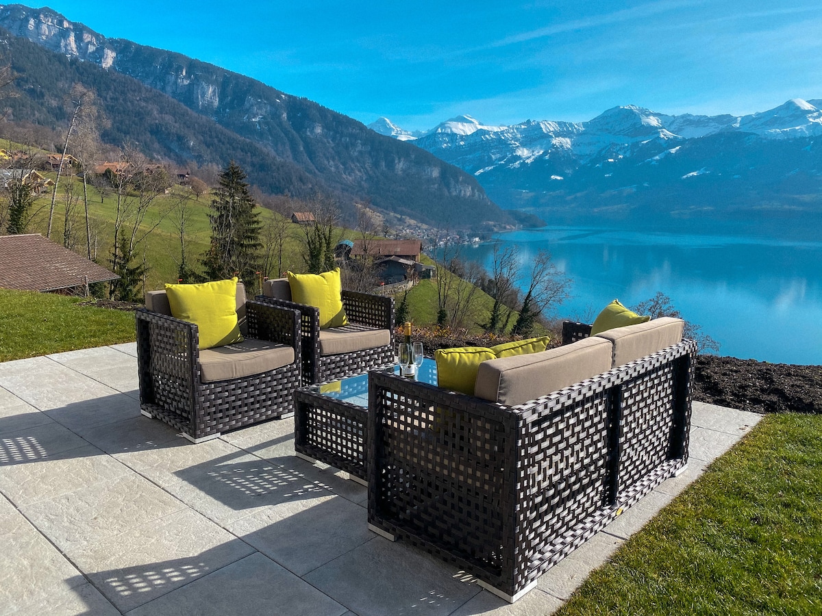 A spacious terrace is furnished with a set of rattan sofas adorned with bright yellow cushions, providing a comfortable seating area. The stunning view features the Thunersee and surrounding mountains, capturing the tranquility of the Sigriswil landscape.