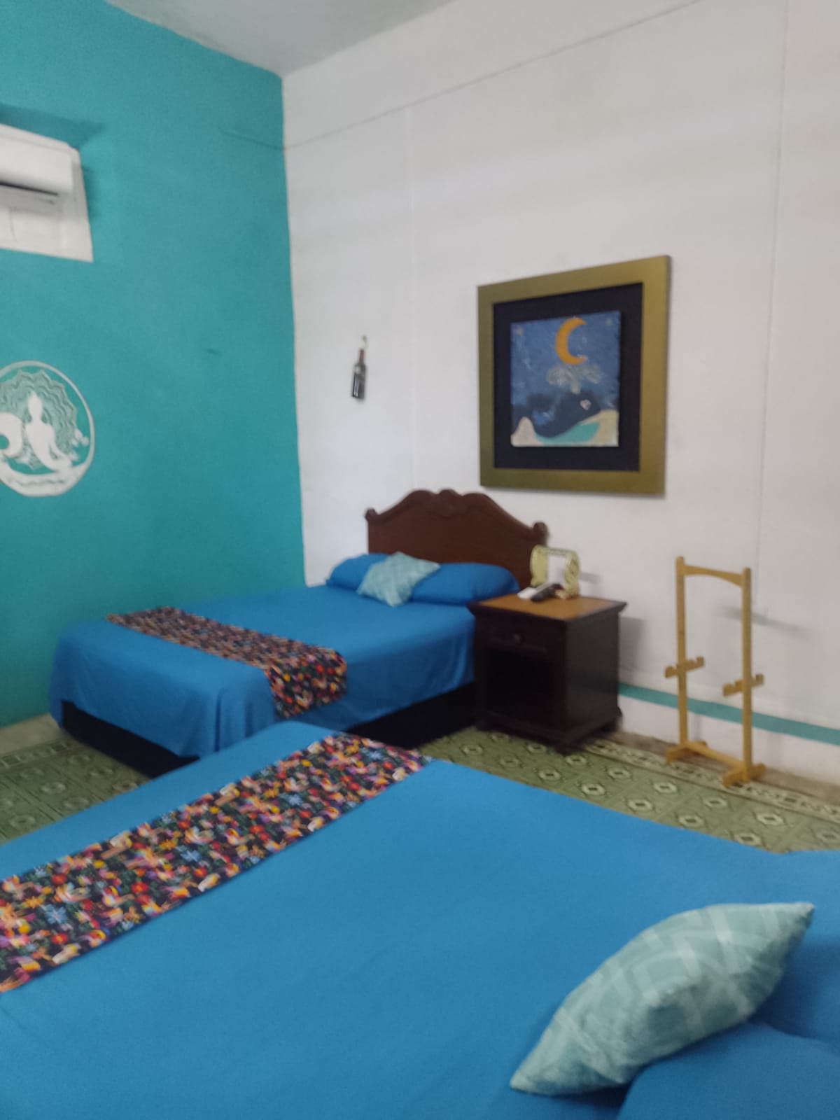 A room featuring two spacious beds with blue covers, complemented by decorative throws. A piece of art depicting a night sky is mounted on a turquoise wall, while a nightstand holds a lamp and other essentials. An air conditioning unit is visible near the ceiling.