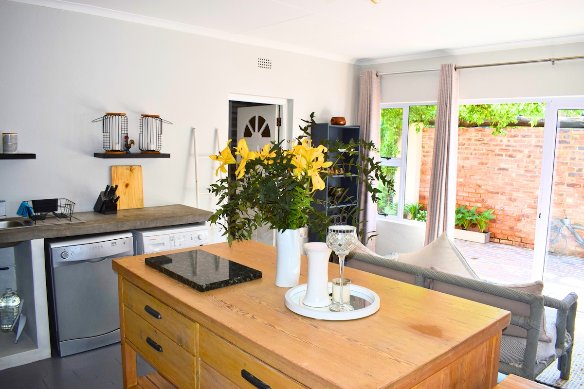The spacious kitchen features a wooden island with a dark countertop, surrounded by a sink and appliances. A vase of yellow flowers adds a touch of color. Natural light streams in through large glass doors, providing views of the private garden beyond.